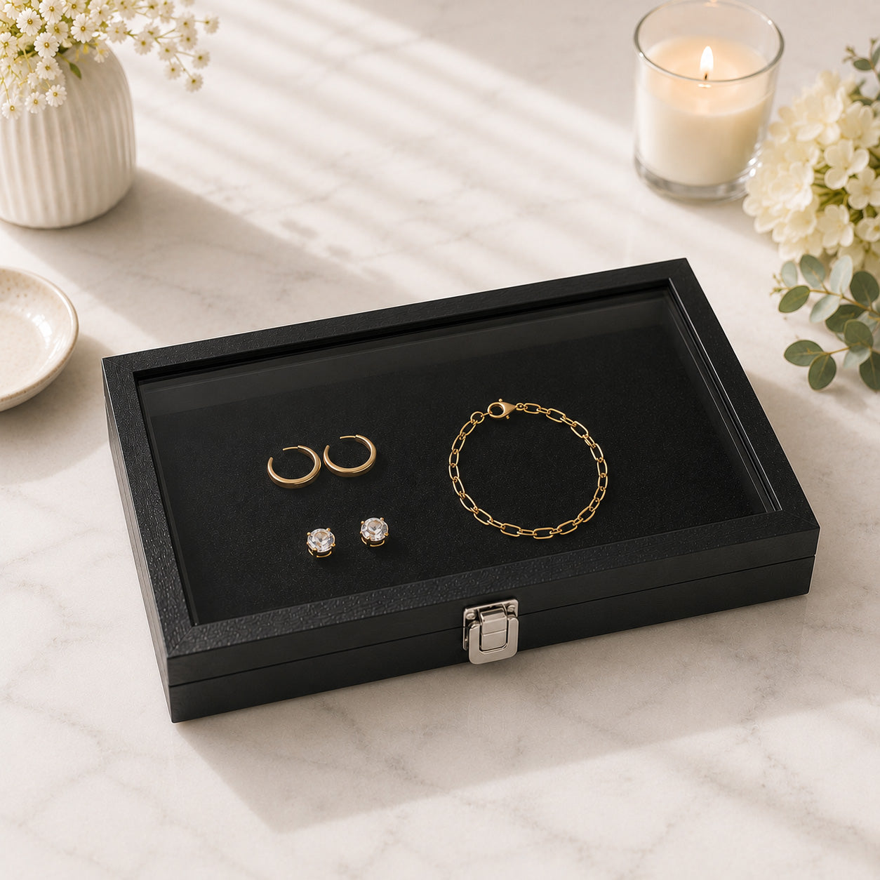 A black jewelry case with a glass lid sits on a light marble surface. Inside the box are gold hoop earrings, a gold chain bracelet, and two small clear stud earrings.