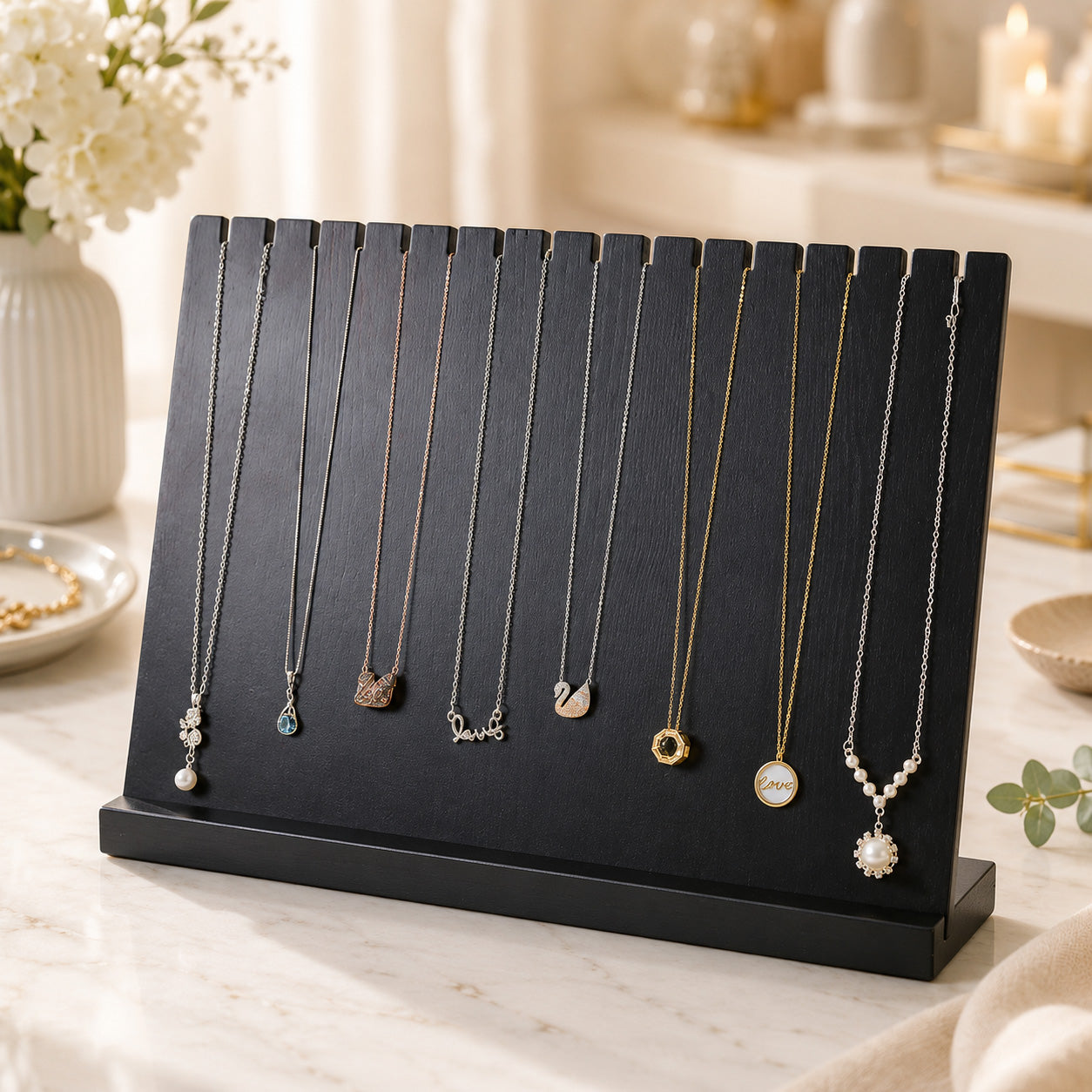 A black wooden necklace display stand holding nine delicate necklaces with various pendants, styled on a marble tabletop with soft-focus decorative props in the background.