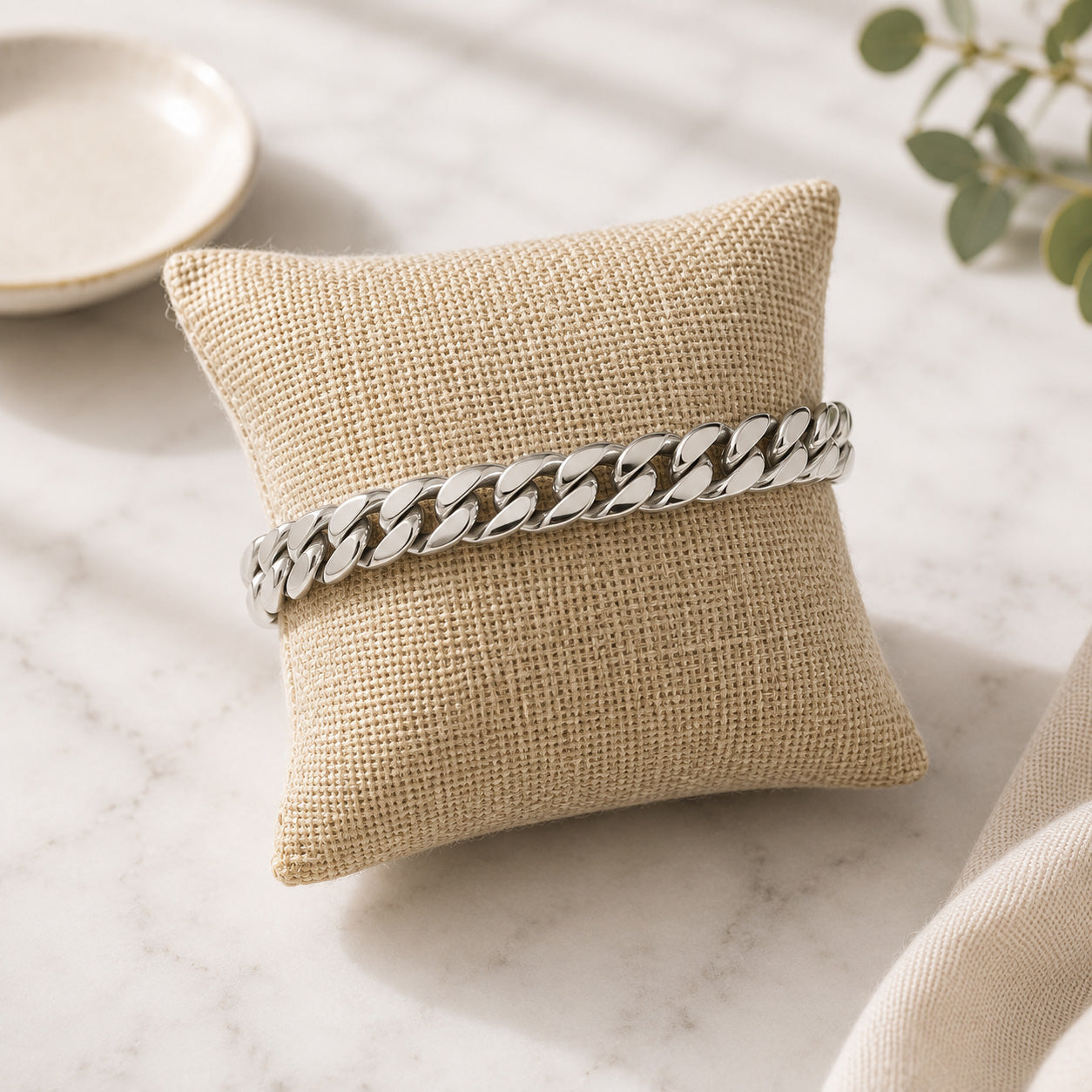 A silver cuban link bracelet on a small beige burlap pillow, set on a light marble surface with soft decorative props.
