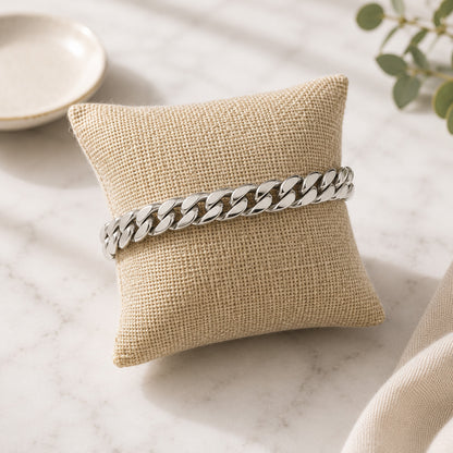 A silver cuban link bracelet on a small beige burlap pillow, set on a light marble surface with soft decorative props.