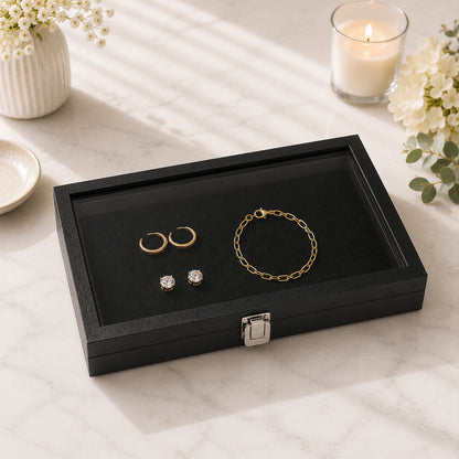 A black jewelry case with a glass lid sits on a light marble surface. Inside the box are gold hoop earrings, a gold chain bracelet, and two small clear stud earrings.